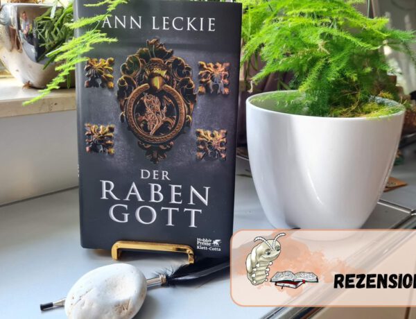 Buch steht auf einem goldenen Buchständer, davor eine schwarze Feder und ein Stein, im Hintergrund Pflanzen